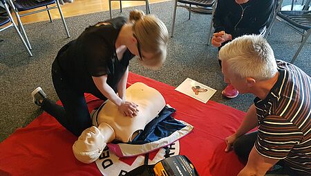 Rehakurs szenische darstellung Erster Hilfe Kurs: Frau kniet vor Puppe und führt Herzdruckmassage durch, ein Mann sitzt kniend daneben