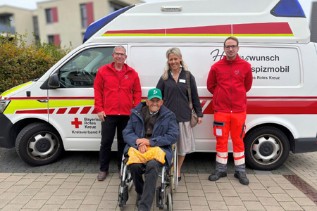Herzenswunschmobil Zwei ehrenamtliche Mitarbeitende des Herzenswunschmobils in ihren roten Einsatzjacken mit Georg Huber, alias der Billige Jakob, im Rollstuhl sitzend und seiner Begleitung. Im Hintergrund das Herzenswunschmobil.