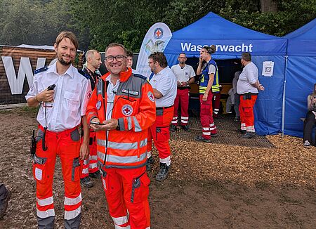 OpenBeatz Zwei Sanitäter lächeln in die Kamera. Im Hintergrund zusehen ist das Festivalgelände des Open Beatz und weitere Sanitäter.