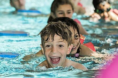 Wasserwacht Schwimmunterricht Foto: Ein Junge lernt mit viel Freude schwimmen. Im Hintergrund sind weitere Kinder im Schwimmbecken zu sehen.